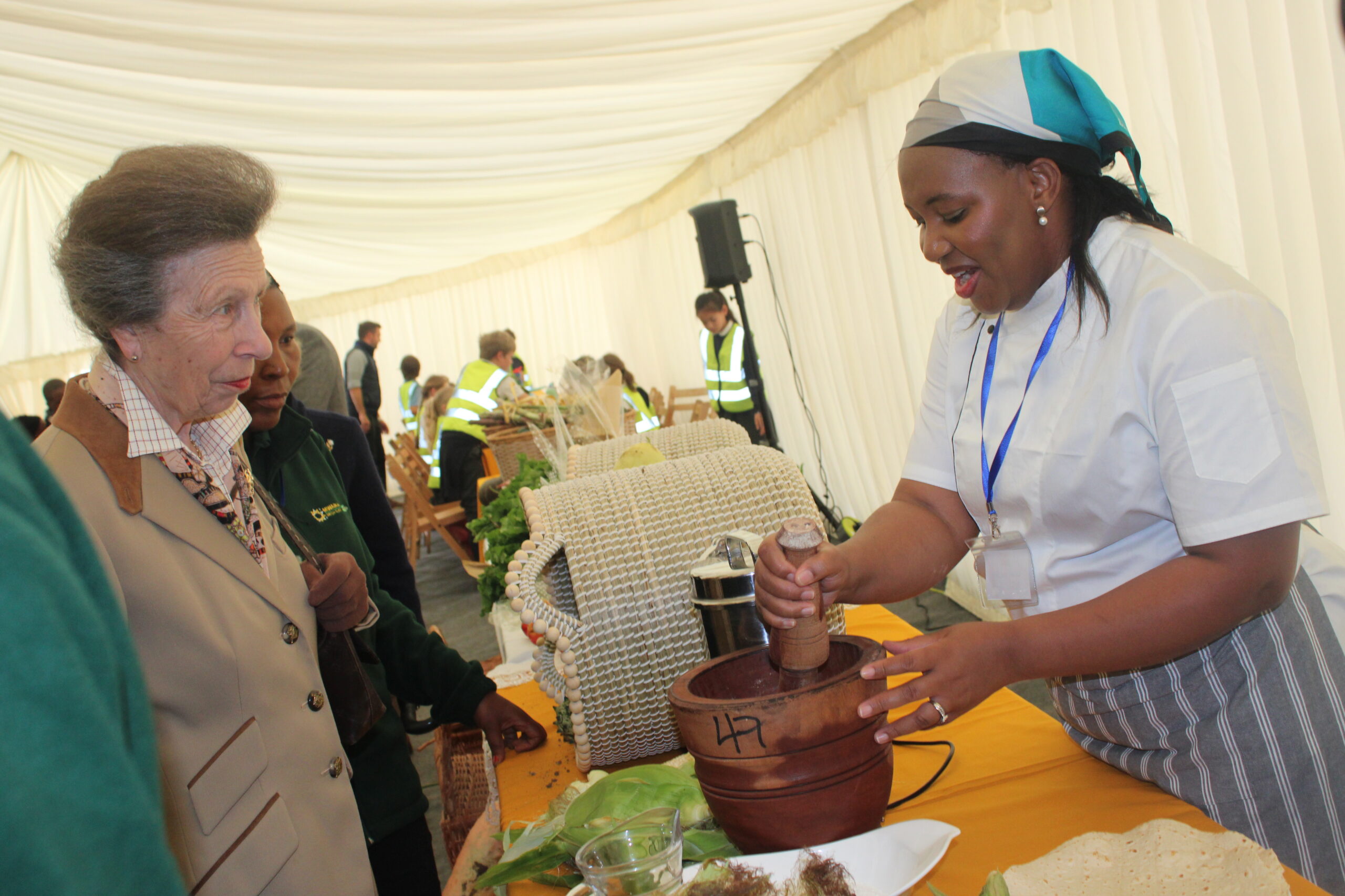 Princess Anne watches a farm worker at Mwanaka Farm in Cambridge demonstrating the traditional process of grinding. maize to prepare sad, showcasing diaspora farming culture.