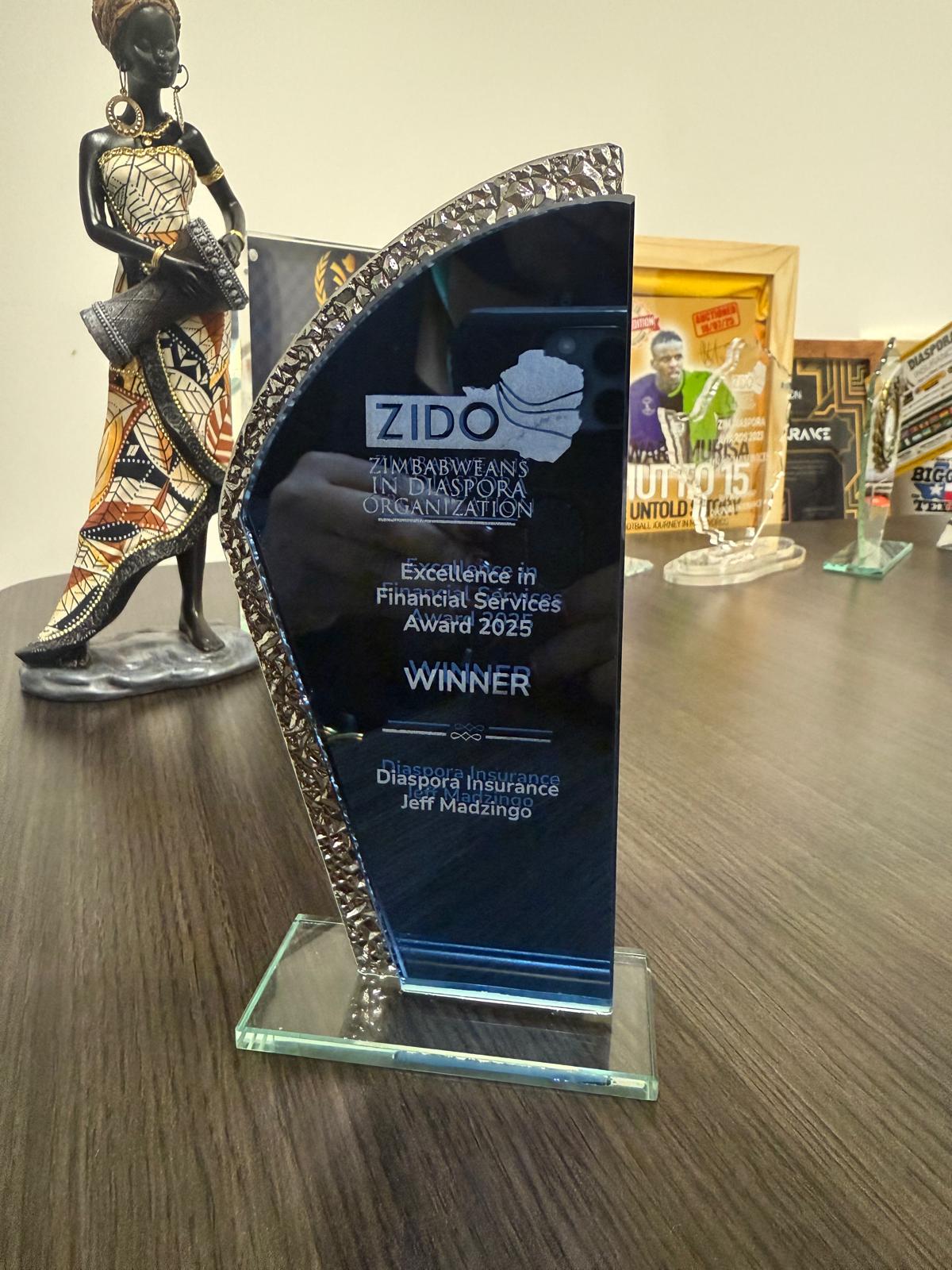 Jeff Madzingo honoured for Empowering the African Diaspora. Glass award trophy engraved with &ldquo;Zimbabweans in Diaspora Organization &ndash; Excellence in Financial Services Award 2025 &ndash; Winner Jeff Madzingo, Diaspora Insurance,&rdquo; displayed on a desk with other accolades.