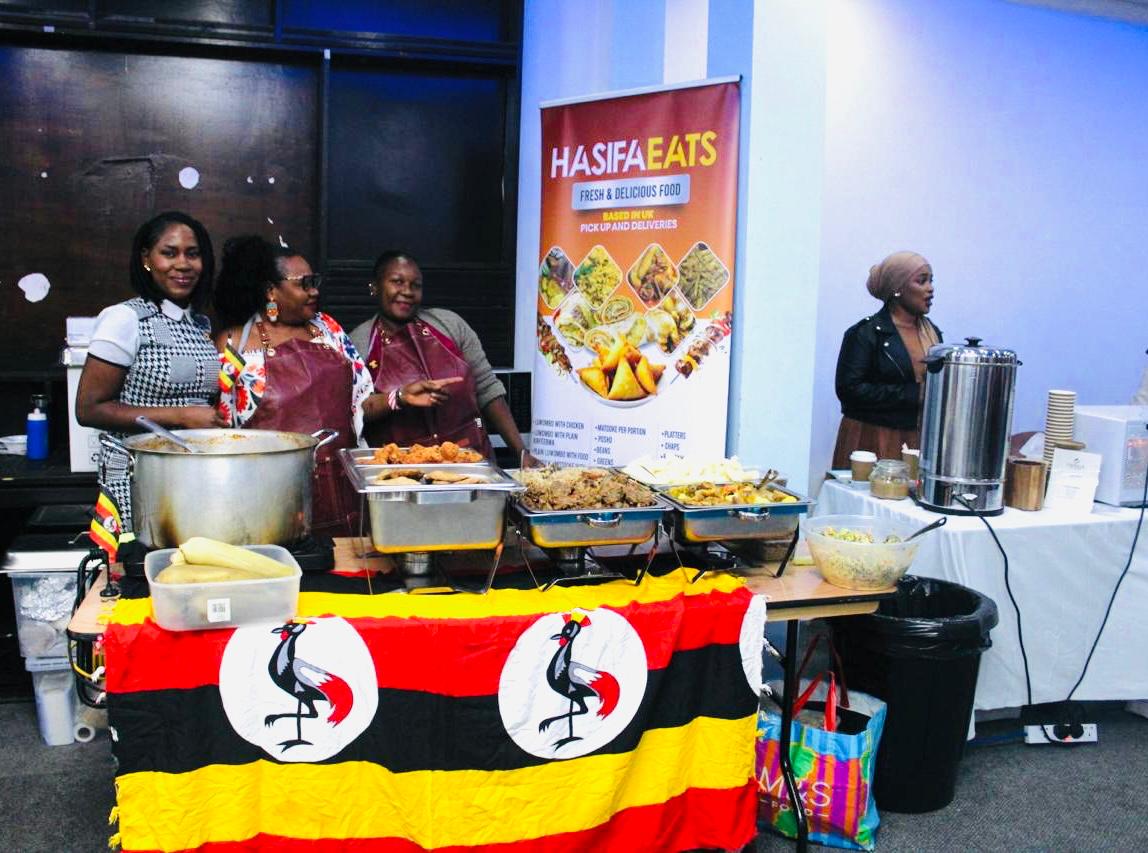Ugandan women serving traditional food and drinks at the Uganda @ 63 Independence celebrations in London, with a Hasifa Eats food stall decorated with the Ugandan flag.