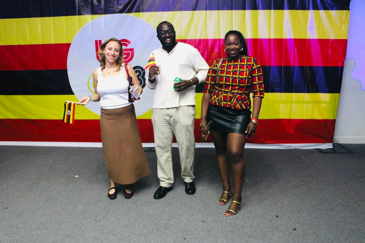 Guests celebrating Uganda @ 63 Independence in London, standing in front of a Ugandan flag backdrop holding national flags and drinks.