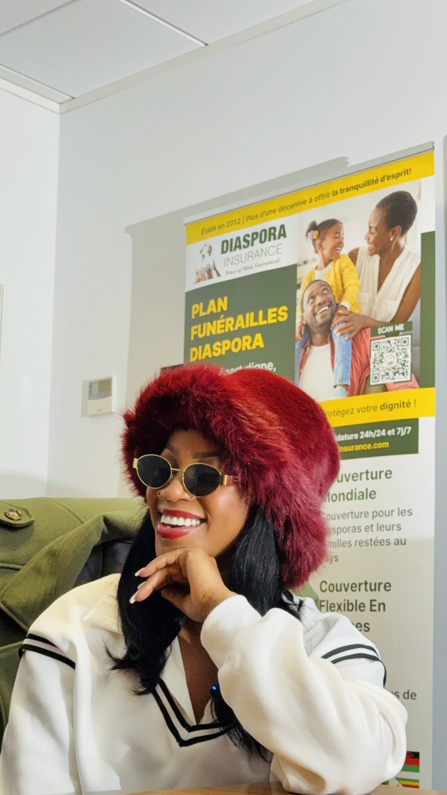 QueenSheebah Karungi wearing a burgundy fur hat, sunglasses, and a white top while seated in the boardroom Diaspora Insurance